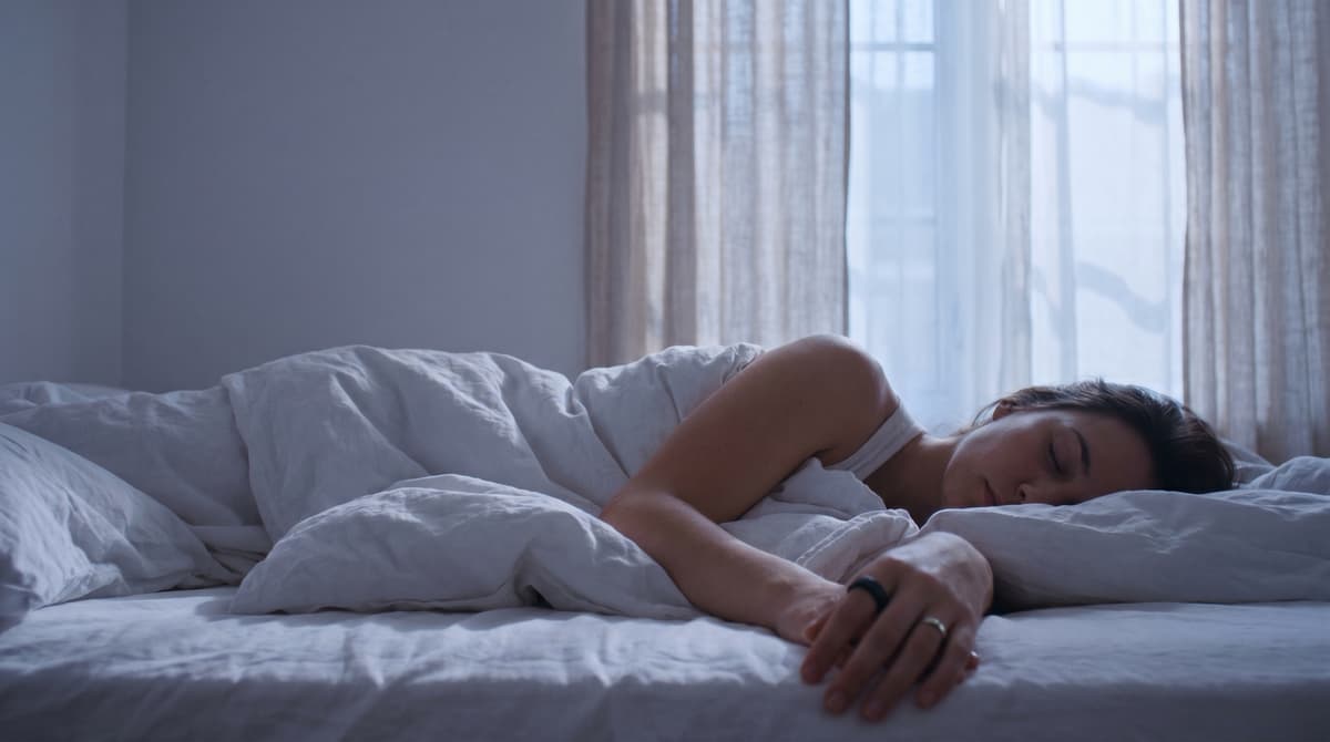 Person sleeping in late morning sunlight in a softly lit bedroom, fitness ring visible on hand resting on white sheets